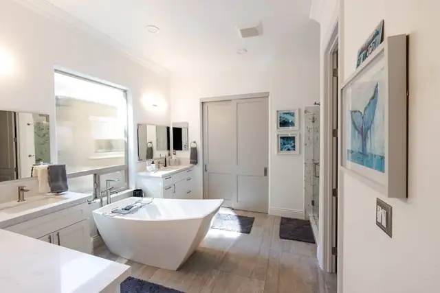 Remodeled primary bathroom with freestanding white tub, wood-look tile floor, white vanities and a large window