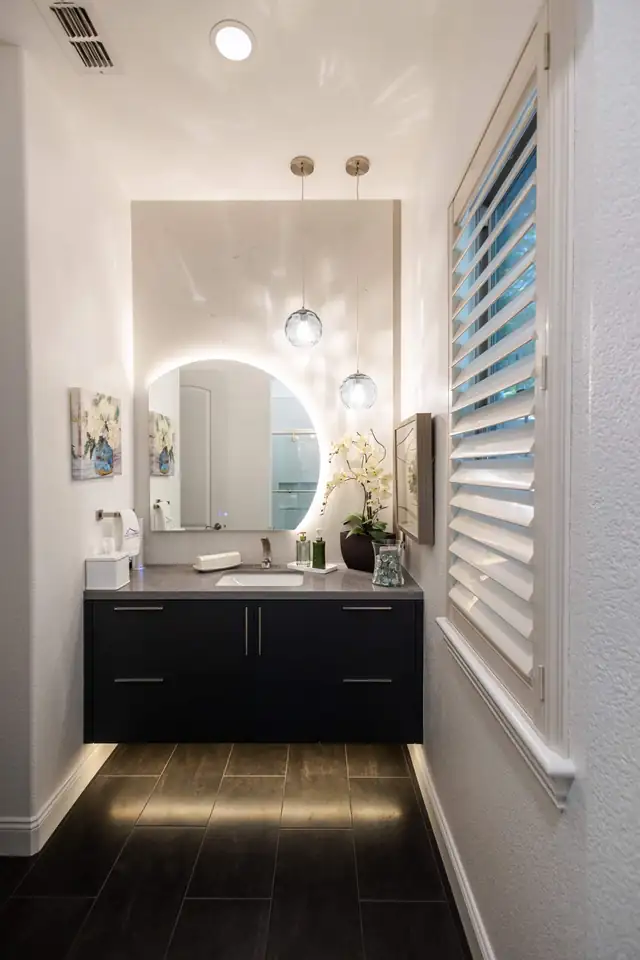 Small bathroom with floating black vanity, under-cabinet lighting, round backlit mirror and glass globe pendants