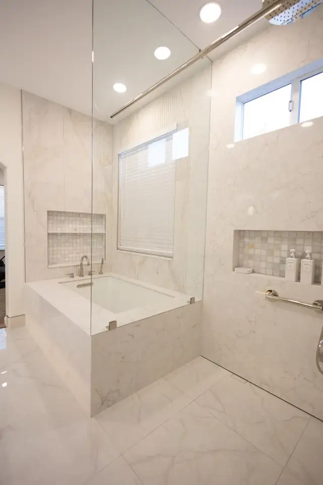 Bathroom with a deep soaking tub and a glass-walled shower in white marble-look tile with recessed niches and a window above